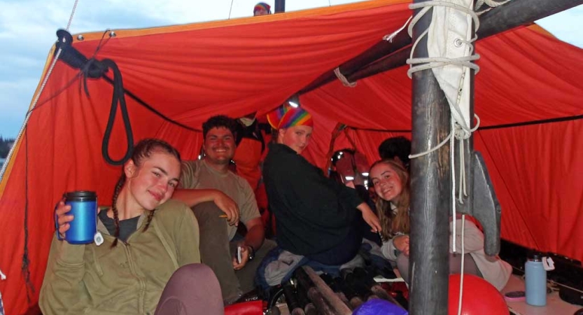 A group of people sit under a tarp shelter set up on a sailboat. 